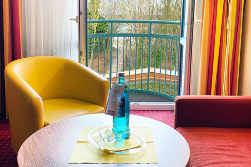 Gemütliches Hotelzimmer mit Balkonblick Hotelzimmer mit gelbem Sessel, rotem Sofa und Tisch vor einem offenen Balkon mit Blick ins Grüne.